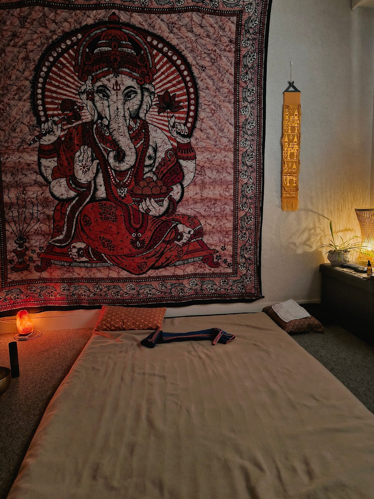 La salle de massage : un futon couvert d'un drap beige posé au sol, une grande tapisserie batik rouge représentant Ganesh, une lampe de sel allumée à gauche et une lampe tressée à droite.